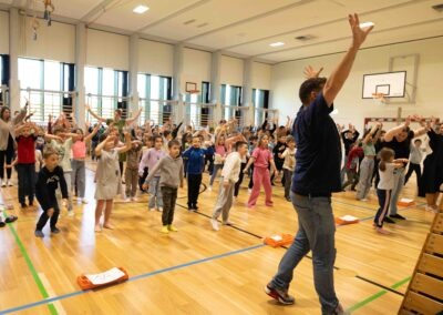 Eine Lehrkraft leitet gemeinsam mit Schülern eine Tanzübung in einer voll besetzten Turnhalle an – Symbol für Inklusion und gemeinsame Bewegung.
