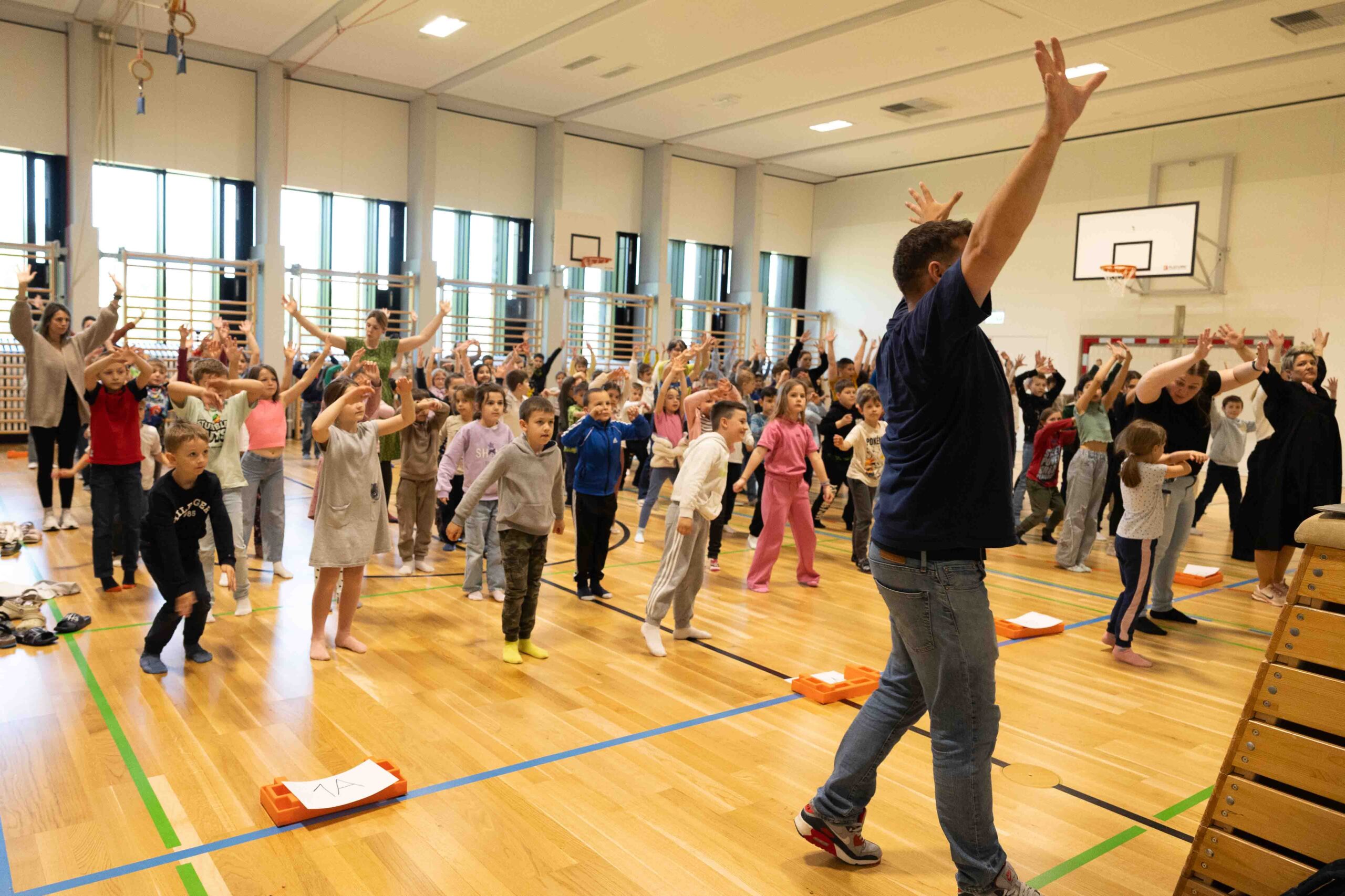 Tanz im Schulalltag: Eine Chance für jedes Kind Eine Lehrkraft leitet gemeinsam mit Schülern eine Tanzübung in einer voll besetzten Turnhalle an – Symbol für Inklusion und gemeinsame Bewegung.