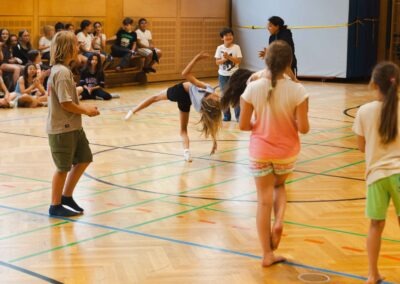 Eine Schülerin macht einen Radschlag in einer Turnhalle, während andere Kinder im Hintergrund zusehen – Symbol für individuelle Ausdruckskraft und Gemeinschaft.