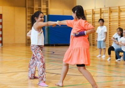 Zwei Schülerinnen in einer Turnhalle bei einer koordinierten Tanzbewegung – inspiriert vom Gemeinschaftsgeist kultureller Tanzbewegungen.
