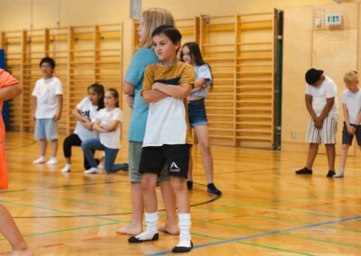 Zwei Schüler stehen Rücken an Rücken mit verschränkten Armen in einer Turnhalle, während im Hintergrund andere Kinder zusehen – Ausdruck von Selbstbewusstsein und Teamgeist.