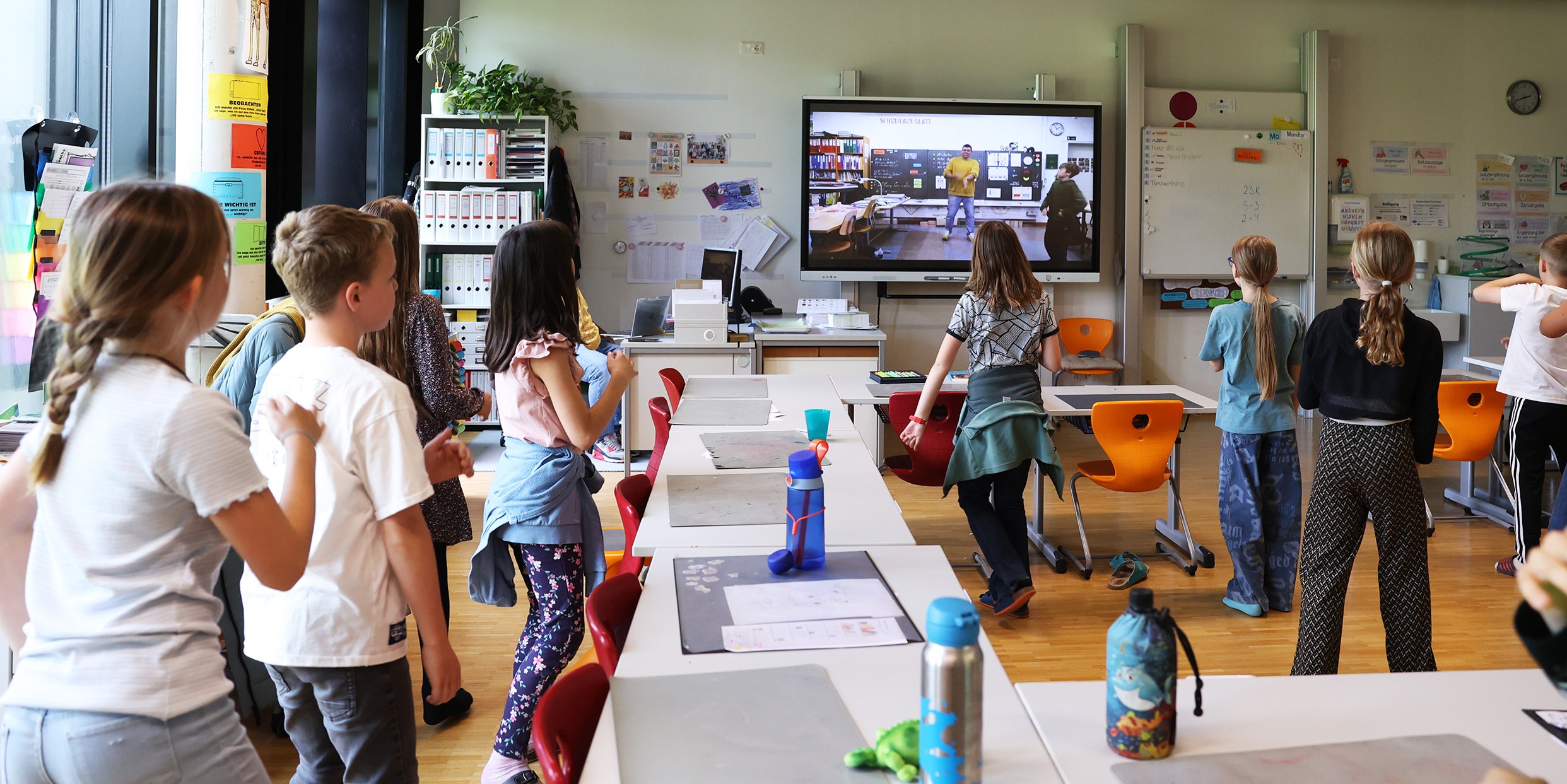 Digitales Unterrichtsmittel für Schulen | groove2grow Eine Schulklasse tanzt und bewegt sich im Klassenzimmer vor einem großen digitalen Bildschirm, auf dem ein groove2grow Video als staatlich geprüftes Unterrichtsmittel läuft.