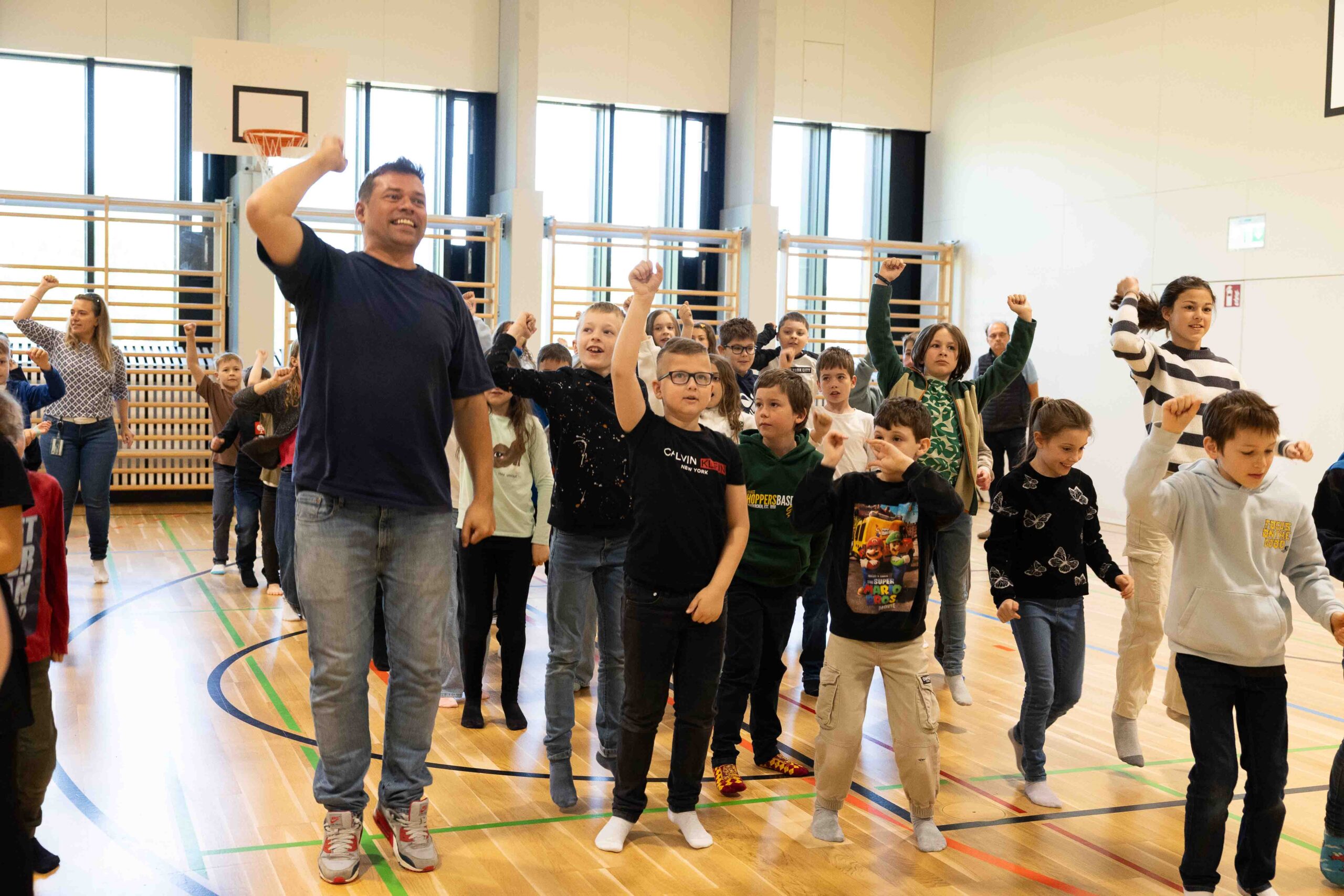 Ein Lehrer tanzt gemeinsam mit einer Gruppe motivierter Schulkinder in einer modernen Turnhalle – praktische Anwendung der groove2grow Lernplattform.