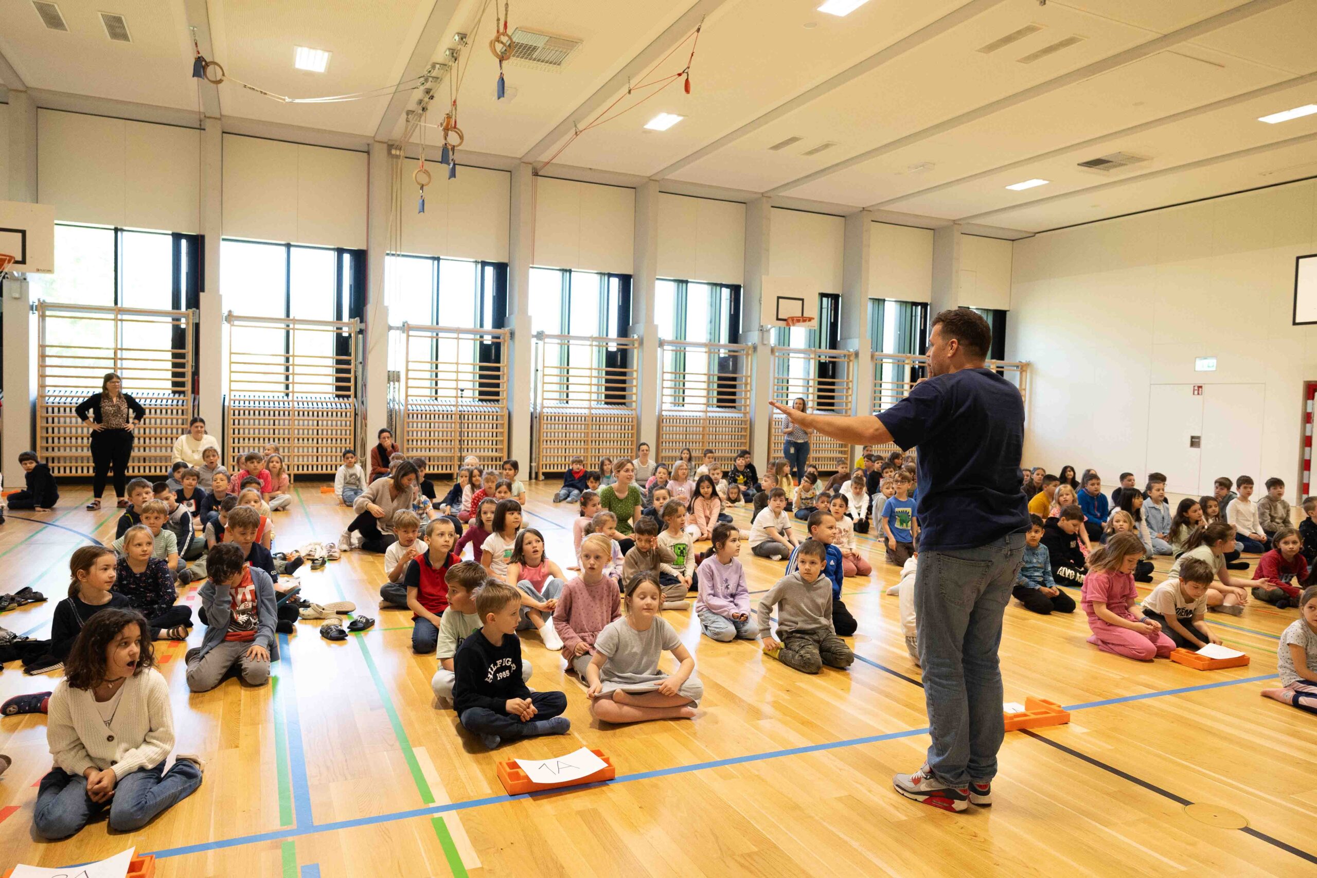 Eine große Gruppe von Schülern sitzt aufmerksam in einer Turnhalle und hört einem Pädagogen zu – Verbindung von Wissenschaft und Schulpraxis.